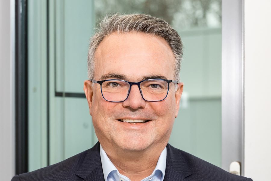 Theis headshot- short hair, glasses, in suit, smiling 